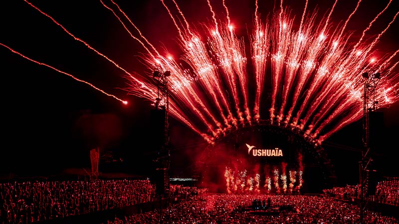 A large crowd watches red fireworks above a stage with the Ushuaïa logo during a nighttime outdoor concert or festival.